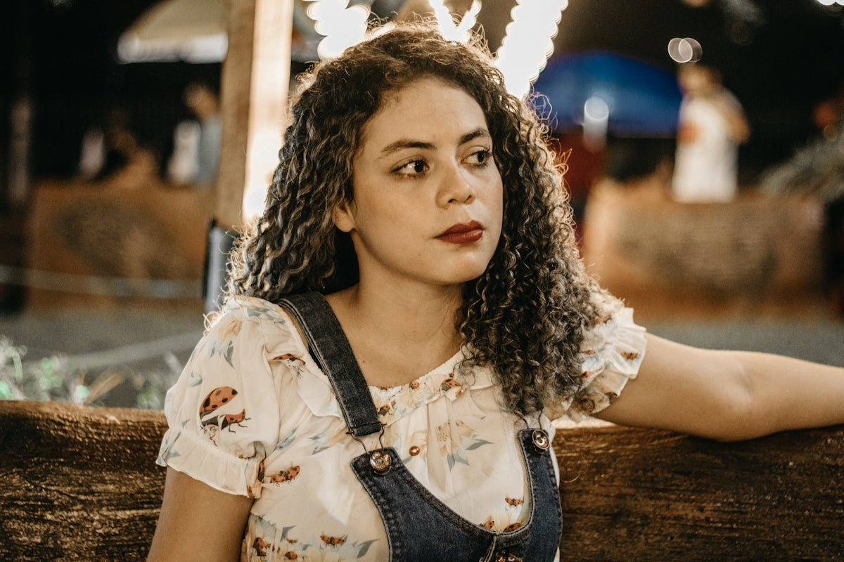 a woman with curly hair wearing overalls leaning on a fence